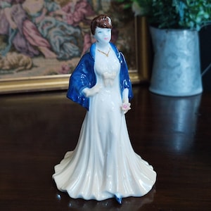 May include: A porcelain figurine of a woman in a long white dress and a blue shawl. The figurine has brown hair and is holding a pink flower. A gold necklace is visible. The figurine is standing on a dark wooden surface.