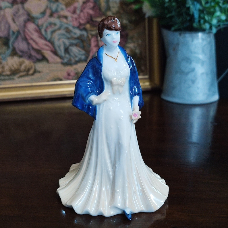 May include: A porcelain figurine of a woman in a long white dress and a blue shawl. The figurine has brown hair and is holding a pink flower. A gold necklace is visible. The figurine is standing on a dark wooden surface.