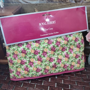 May include: A floral tea cosy with a green background and a pattern of pink and yellow roses. The tea cosy is trimmed with a pink border. The packaging reads "ROYAL ALBERT Tea Cosy Coordinated Kitchen Textiles."