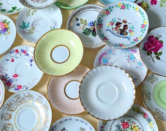 May include: A collection of assorted ceramic saucers featuring various designs, including floral patterns, gold-trimmed edges, solid pastel colours, and a commemorative plate with a portrait of Queen Elizabeth II and Prince Philip dated 1952-1977.
