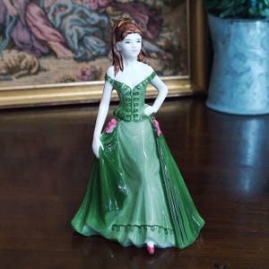 May include: A porcelain figurine of a woman in a green gown with pink accents. The figurine has brown hair and is posed with one hand on her hip. The dress has a detailed bodice and a flowing skirt. The figurine is a decorative item.