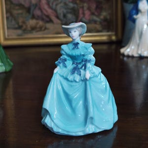 May include: A porcelain figurine of a woman in a long, light blue dress and a matching hat. The dress features dark blue bows and ruffles. The figurine is a decorative item, likely for display.