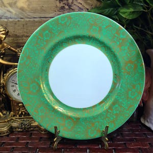 May include: A round ceramic plate featuring a solid white centre surrounded by a wide, vibrant green border. The green border is decorated with an intricate, repeating gold floral and lace-like pattern that covers the entire rim area.