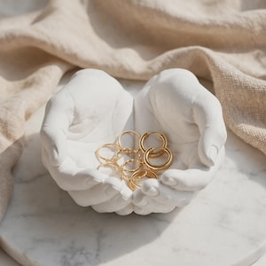 May include: A white, matte-finished sculpture shaped like two cupped hands holding a collection of gold-toned metal rings and hoop earrings. The sculpture features realistic finger and palm details, providing a concave surface for small items.