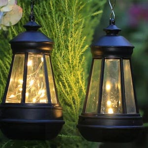 Puede incluir: Dos faroles colgantes negros con paneles de vidrio transparente y forma cónica. Cada farol cuenta con un marco de metal negro y un asa superior para colgar. En el interior, luces de cadena de color blanco cálido iluminan el espacio.