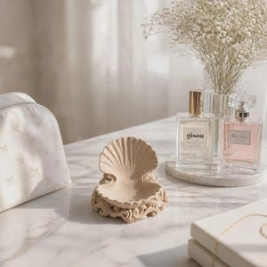 Peut inclure: Un vide-poche beige en forme de coquillage avec des volutes ornementales à la base. L'objet est placé près d'une trousse de toilette blanche à motifs de nœuds, deux flacons de parfum en verre et une pile de livres blancs sur une surface en marbre.