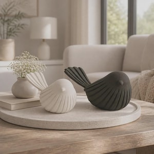 Peut inclure: Deux figurines d'oiseaux, une en blanc mat et une en noir mat, présentant un design stylisé avec des détails nervurés verticaux sur les ailes et le corps. Les oiseaux sont placés côte à côte sur un plateau circulaire en pierre claire.
