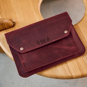 Può includere: Una custodia per laptop in pelle bordeaux con chiusura a patta fissata con due bottoni automatici in metallo. La custodia presenta la lettera "W" con un design a corona d'alloro. La custodia è su un tavolo di legno.