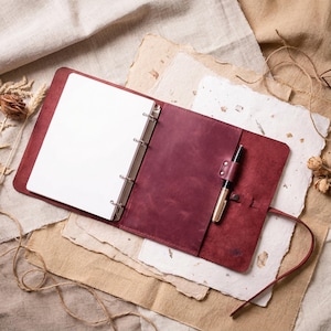 Puede incluir: Carpeta de cuero burdeos abierta, que revela papel blanco en blanco, con un bolígrafo asegurado en un lazo de cuero. La carpeta descansa sobre una superficie texturizada con flores secas y cuerda.