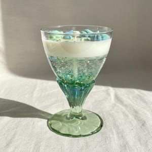 May include: A decorative candle in a stemmed glass. The candle has a white top layer and a lower layer with green glitter and bubbles. The glass has a green stem and base. The top of the candle is decorated with blue and white seashell-shaped wax pieces.