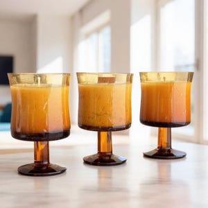 May include: Three amber-colored glass candle holders with a stem and base, each filled with a light orange wax. The candle holders are arranged on a white surface, with a bright background.