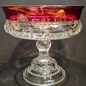 May include: A clear glass pedestal bowl with a ruby red rim. The bowl has a decorative pattern of circles and a detailed stem. The base is round and also features a decorative pattern. The bowl is likely used for serving desserts or displaying decorative items.