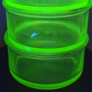 May include: Two stacked, circular, translucent neon green glass containers. The glass exhibits a bright, glowing appearance, characteristic of uranium glass under ultraviolet light, featuring smooth, rounded edges and a simple, cylindrical design.