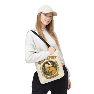 May include: Cream-colored tote bag featuring the Hufflepuff house crest. The crest includes a badger illustration in a circular design with the words "Hufflepuff" and "Loyal, Patient, True". The bag has a black shoulder strap.