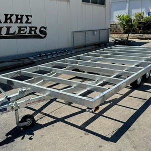 Op de afbeelding: Zilveren metalen aanhangerframe met een rechthoekig ontwerp, voorzien van een trekhaak, wielen en een steunstandaard. De aanhanger staat op een betonnen ondergrond, met de bedrijfsnaam "ΛΙΤΣΑΚΗΣ TRAILERS" zichtbaar.