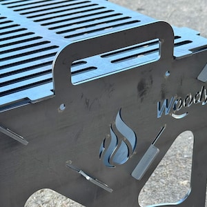 May include: A metal portable grill with a slotted cooking surface and a handle. The side panel features a flame design, the word "Woody", and cutouts of grilling tools. The grill is made of a dark gray metal.