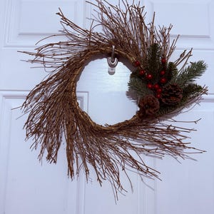 May include: A rustic wreath made of woven brown twigs, adorned with pine needles, red berries, and pine cones. The wreath is circular and hangs on a white door, creating a natural and festive decoration.