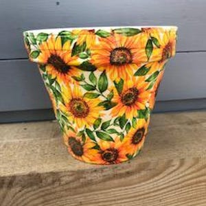 May include: A tapered plant pot featuring a vibrant all-over pattern of yellow and orange sunflowers with green leaves against a white background. The pot has a wide, flared rim and a smooth, glossy finish, showcasing multiple sunflower blooms.