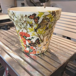 May include: A tapered ceramic plant pot featuring a decorative decoupage pattern of orange flowers, green foliage, yellow blossoms, butterflies, and small birds against a cream-coloured background with a smooth, glossy finish and a wide rim.