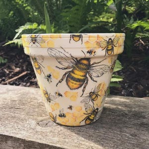 May include: A cream-coloured terracotta plant pot featuring a repeating pattern of yellow and black honeybees and golden honeycomb hexagonal shapes. The design covers the exterior surface of the pot, including the rim, with various bee sizes.