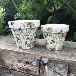 May include: Two white ceramic flower pots of different sizes, both featuring a botanical pattern of daisies, green foliage, and small bees. The pots have a tapered shape with a wide rim and a smooth finish, displaying a consistent floral design.