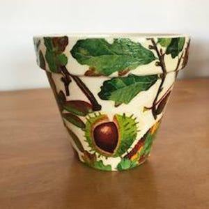May include: A cream-coloured ceramic plant pot featuring a detailed botanical pattern of green leaves, brown twigs, and spiky green husks containing dark brown chestnuts. The design wraps around the exterior of the tapered, wide-rimmed vessel.