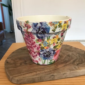 May include: A tapered ceramic plant pot featuring a vibrant decoupage floral pattern. The exterior is decorated with watercolour-style illustrations of pink, blue, and yellow flowers against a white background, with a plain cream-coloured interior rim.