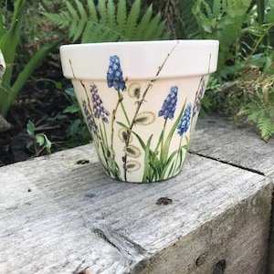 May include: A cream-coloured ceramic flower pot featuring a hand-painted botanical design. The exterior displays blue grape hyacinth flowers, green stems, and fuzzy pussy willow branches arranged vertically around the tapered cylindrical body.