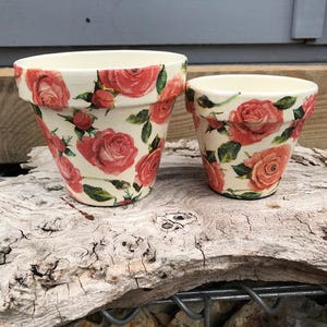 May include: Two cream-coloured ceramic flower pots of different sizes, both decorated with a repeating pattern of pink roses, green leaves, and small buds. The pots feature a wide, flared rim and a tapered body, showcasing a floral design.
