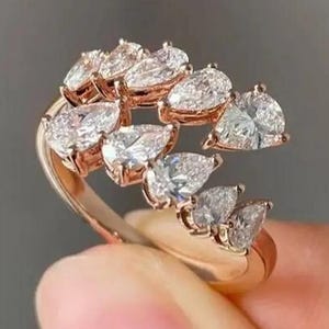 May include: A rose gold ring with multiple clear, pear-shaped gemstones. The gemstones are arranged in a flowing, asymmetrical design. The ring is held up against a neutral background.