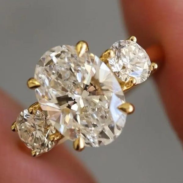 May include: A gold ring with a large oval-cut diamond and two smaller round diamonds. The ring's setting is a classic design, highlighting the sparkle of the clear gemstones. The ring is held up by a finger.