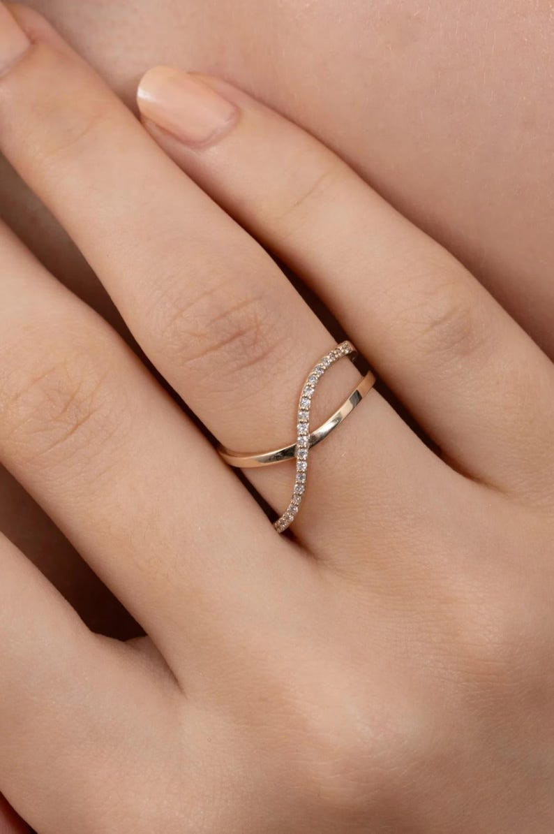 Puede incluir: Un anillo dorado con un dise&ntilde;o &uacute;nico e entrelazado. Una banda est&aacute; adornada con peque&ntilde;as piedras brillantes, creando un aspecto delicado y elegante. El anillo se muestra en un dedo, resaltando su forma y detalles.