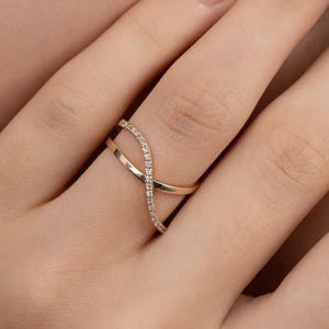 Puede incluir: Un anillo dorado con un dise&ntilde;o &uacute;nico e entrelazado. Una banda cruza otra, con una banda engastada con peque&ntilde;as piedras brillantes. El anillo se muestra en el dedo de una persona.