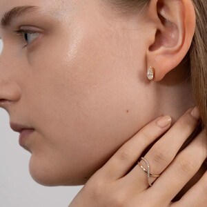 Puede incluir: Primer plano de una oreja con un peque&ntilde;o pendiente de oro con un diamante en forma de l&aacute;grima. Un anillo de oro con un dise&ntilde;o entrecruzado y peque&ntilde;os diamantes est&aacute; en el dedo de la persona. La joyer&iacute;a est&aacute; sobre un fondo neutro.