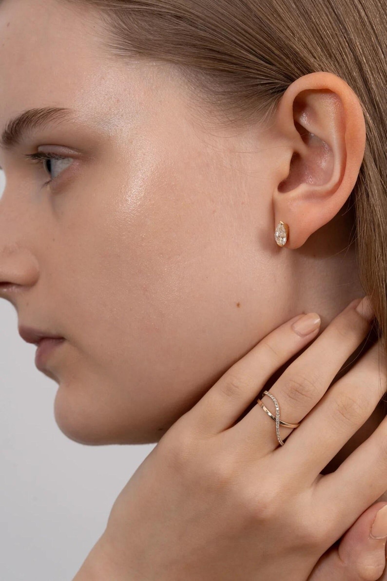 Puede incluir: Primer plano de una oreja con un peque&ntilde;o pendiente de oro con un diamante en forma de l&aacute;grima. Un anillo de oro con un dise&ntilde;o entrecruzado y peque&ntilde;os diamantes est&aacute; en el dedo de la persona. La joyer&iacute;a est&aacute; sobre un fondo neutro.