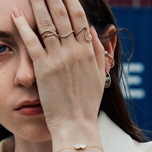 Puede incluir: Primer plano de una mano adornada con tres anillos de oro, cada uno con un dise&ntilde;o &uacute;nico y adornado con peque&ntilde;as piedras brillantes. Tambi&eacute;n se ve una delicada pulsera de oro con una gema rectangular. Las joyas se destacan sobre un fondo suave.