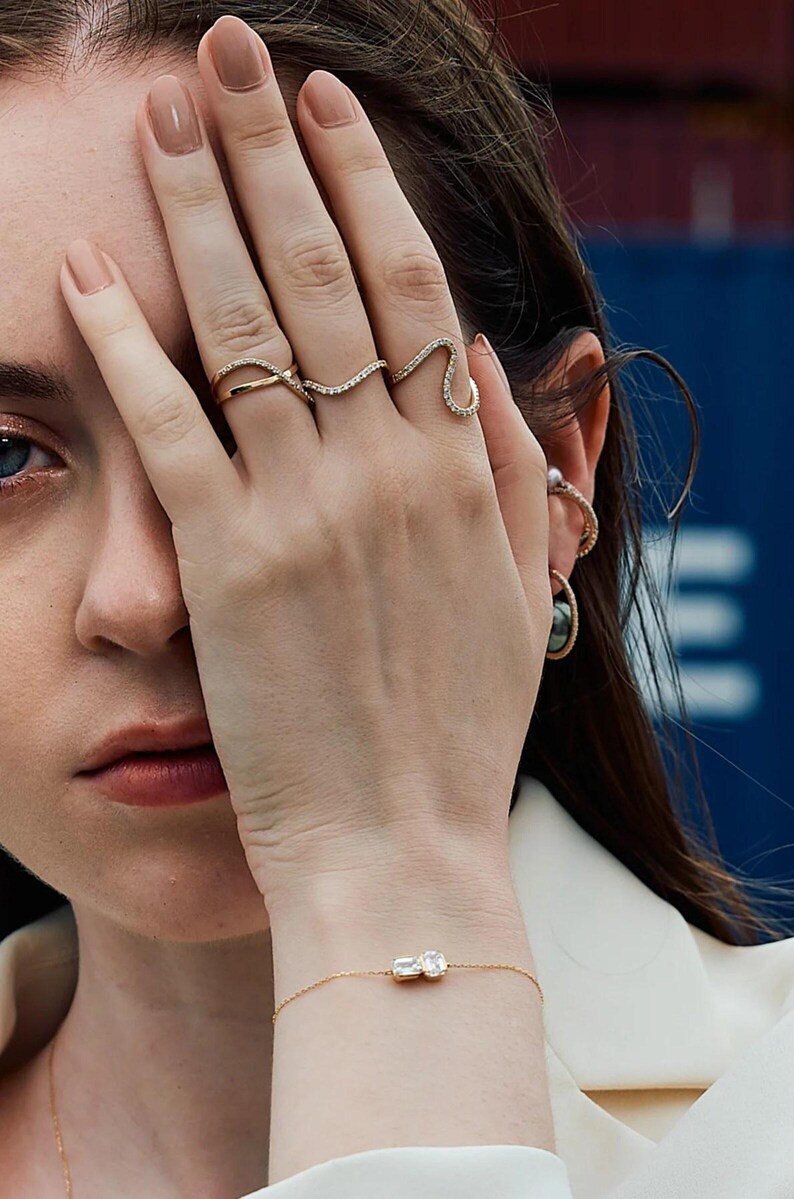 Puede incluir: Primer plano de una mano adornada con tres anillos de oro, cada uno con un dise&ntilde;o &uacute;nico y adornado con peque&ntilde;as piedras brillantes. Tambi&eacute;n se ve una delicada pulsera de oro con una gema rectangular. Las joyas se destacan sobre un fondo suave.