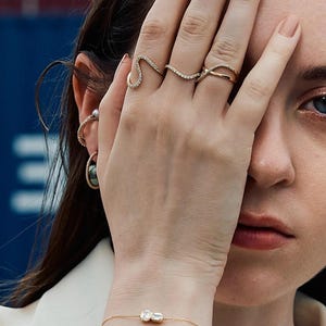 Puede incluir: Una mano adornada con anillos de oro, incluyendo un anillo en forma de serpiente y una banda delicada. Una pulsera de oro con una gema rectangular es visible en la mu&ntilde;eca. La imagen muestra joyer&iacute;a fina, enfatizando la elegancia y el dise&ntilde;o de las piezas.