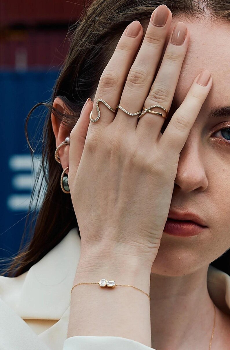 Puede incluir: Una mano adornada con anillos de oro, incluyendo un anillo en forma de serpiente y una banda delicada. Una pulsera de oro con una gema rectangular es visible en la mu&ntilde;eca. La imagen muestra joyer&iacute;a fina, enfatizando la elegancia y el dise&ntilde;o de las piezas.