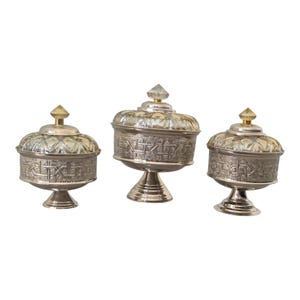 Op de afbeelding: Drie zilverkleurige decoratieve kommen met deksels. Elke kom heeft een voet en een gedetailleerd, patroonontwerp op de body en het deksel. De deksels zijn bekroond met een kristallen of goudkleurige finial.