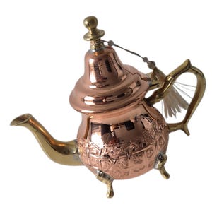Puede incluir: Una tetera de color cobre con boquilla, asa y detalles dorados. La tetera tiene un patrón decorativo en el cuerpo y una pequeña borla unida a la tapa. Descansa sobre tres patas pequeñas.