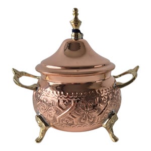 Op de afbeelding: Een koperkleurige suikerpot met deksel, versierd met bloemmotieven en messing handvatten en poten. Het deksel is bekroond met een decoratieve eindknop. De body van de kom heeft gedetailleerde reliëfontwerpen, wat bijdraagt aan de vintage esthetiek.
