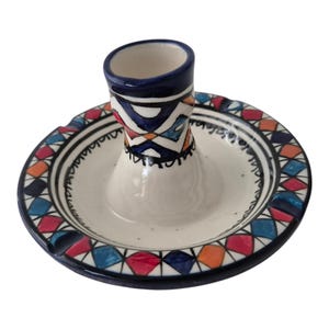 Op de afbeelding: Een keramische asbak met een centrale cilindrische houder en een brede, cirkelvormige basis. De asbak is voornamelijk wit met een blauwe rand en kleurrijke geometrische patronen in rood, blauw, oranje en zwart. Het ontwerp is handgeschilderd.