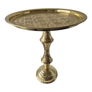 Pode incluir: Uma mesa lateral dourada e ornamentada, com um tampo redondo e estampado. A mesa tem uma base decorativa em estilo pedestal. A mesa de metal é provavelmente feita de latão ou material semelhante. A mesa é adequada para exibir itens decorativos.