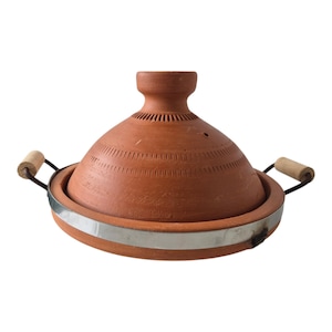 Op de afbeelding: Een terracotta tajine kookpot met een conisch deksel voorzien van gegraveerde lineaire patronen en een knopvormige handgreep. De basis is omringd door een zilverkleurige metalen band en heeft twee zijhandgrepen met lichtgekleurde houten grepen.