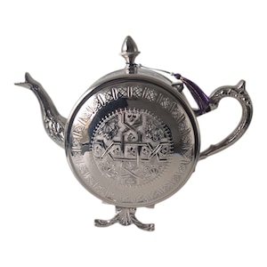 Op de afbeelding: Zilverkleurige theepot met ingewikkelde reliëfpatronen. De theepot heeft een gebogen tuit, een decoratieve handgreep en een kleine paarse kwast. Het ontwerp omvat bloemen en geometrische motieven en de theepot staat op een decoratieve voet.