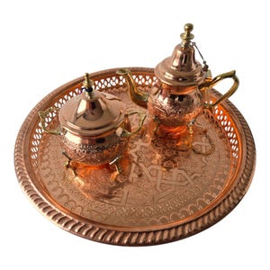 May include: A copper tea set with intricate designs, including a teapot, sugar bowl, and serving tray. The tray has a decorative edge and a detailed pattern in the center. The teapot and sugar bowl have ornate lids and handles.