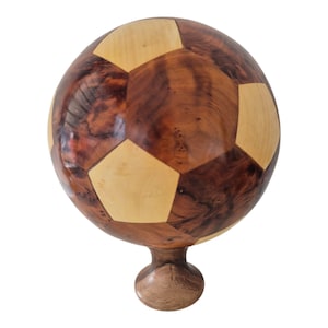 Könnte beinhalten: Ein dekoratives Objekt in Form eines Fußballs aus Holz. Der Ball besteht aus dunkelbraunem und hellbraunem Holz mit einem Muster aus Fünfecken. Er steht auf einem kurzen, abgerundeten Holzsockel. Das Objekt ist einem Fußball nachempfunden.