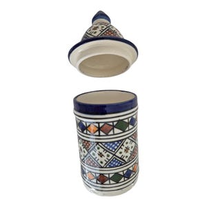 Op de afbeelding: Een cilindrische keramische pot met een bijpassend conisch deksel, voorzien van een handgeschilderd geometrisch patroon in blauw, groen, oranje en bruin op een witte achtergrond. De pot heeft een donkerblauwe rand en het deksel zweeft boven de opening.
