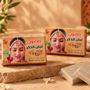 Può includere: Due scatole di sapone rettangolari con l'illustrazione di una persona che indossa gioielli d'oro tradizionali e un anello al naso. La confezione include testo in arabo, un'immagine di resina e un logo circolare verde. Sono visibili due saponette.
